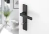 A black door handle with a key inserted into the lock, mounted on a white door. The background shows a blurred interior of a room with furniture and plants. Produktbild-Vorschau