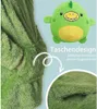 Nahaufnahme eines grünen Plüsch-Decken-Hoodies mit einer Tasche, im Hintergrund ein kleines grünes Dinosaurier-Plüschtier Podgląd zdjęcia produktu