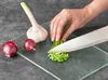 A hand is chopping chives on a glass cutting board next to a head of garlic and two red onions. Produktbild-Vorschau