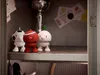 Three small collectible figures are displayed on a shelf inside a locker, with a trophy and some papers in the background. Produktbild-Vorschau
