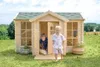 Two children playing in and around a wooden playhouse in a grassy field. Produktbild-Vorschau