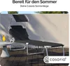A black sun lounger with a canopy is set up on a wooden deck next to a small round table with a drink on it, with a textured wall in the background. Produktbild-Vorschau