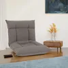 A grey floor chair is shown in a room with a wooden floor and a blue rug. A small wooden side table with a vase of dried flowers is next to the chair. A painting hangs on the wall in the background. Produktbild-Vorschau