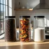 Three glass storage jars with black lids are displayed on a wooden surface in a kitchen setting. Produktbild-Vorschau
