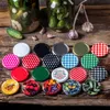 A collection of colorful jar lids arranged on a wooden surface, with jars of pickles and garlic in the background. Voir les images du produit