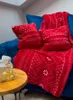 A red blanket and pillows with a white pattern are arranged on a blue sofa, with a small wooden table and a lit gift box in the foreground. Produktbild-Vorschau