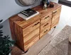 A wooden sideboard with drawers and books on top, placed in a room with a rug and a plant. Produktbild-Vorschau