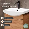 A close-up view of a white ceramic washbasin with a black faucet, mounted on a wooden vanity cabinet. The background is a textured gray wall. Produktbild-Vorschau