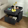 A black nightstand with drawers and open shelves is placed next to a sofa. The top surface has a wireless charger with a phone, a vase with flowers, and a bowl of fruit. Charging cables and adapters are also visible. Podgląd zdjęcia produktu