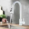 A white kitchen faucet with a pull-out spray head is shown in use over a stainless steel sink. A hand is holding the spray head, demonstrating its functionality. The background includes a gray tiled wall and a wooden countertop with a cutting board and a p Náhľad obrázku produktu