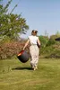 A woman walks away from the camera in a garden, carrying a large dark gray planter with an orange interior and a bunch of green plants. Náhľad obrázku produktu
