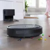 A robot vacuum cleaner is shown on a wooden floor with colorful candies scattered around it. A person in pink pants is walking in the background. Voir les images du produit