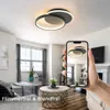 A hand holding a smartphone displaying a ceiling light in a living room setting. The living room has a sofa, coffee table, and windows. Produktbild-Vorschau