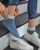 A person is putting on a pair of blue and white socks while wearing jeans and white sneakers, standing on a set of stairs. Produktbild-Vorschau