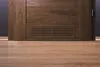 A close-up view of a wooden door with a vent near the bottom, set against a wooden floor. Podgląd zdjęcia produktu