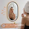 A woman is applying makeup in front of a lighted oval mirror in a bathroom setting. The mirror has an anti-fog feature indicated by arrows and text. Produktbild-Vorschau