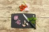 A black cutting board with chopped red onion, garlic cloves, a knife, and parsley, with cherry tomatoes and a garlic bulb on a distressed wooden surface. Produktbild-Vorschau
