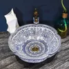 A decorative blue and white patterned basin is shown on a dark marble countertop, with a faucet and a vase in the background. Náhled obrázku produktu