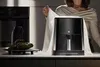 A person is partially visible in the background, holding a white cloth over a black air fryer on a kitchen counter. Produktbild-Vorschau