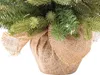 A close-up view of the base of a small artificial Christmas tree, showing its green branches and a burlap pot cover. Voir les images du produit