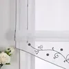 A close-up of a white sheer curtain with a black floral embroidery pattern and a gray trim. A bouquet of white roses is visible on the left side. Produktbild-Vorschau