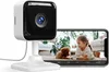 A white indoor security camera is shown next to a smartphone displaying a live feed of a child and a dog. Produktbild-Vorschau