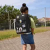 A young girl wearing a backpack stands outdoors on a paved area with a grassy field and buildings in the background. Anteprima dell’immagine prodotto
