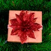 A close-up view of a Christmas gift box decorated with a red glitter poinsettia flower, nestled amongst green pine needles. Produktbild-Vorschau