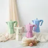 Four colorful Moka pots are arranged on a surface with draped fabric and some dried flowers. Anteprima dell’immagine prodotto