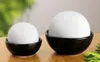 Two white spheres are placed in black bowls on a wooden surface, with a blurred green background. Produktbild-Vorschau
