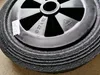 A close-up view of a black wheel with a textured rubber tire and a plastic rim. There is a white sticker with writing on the left side of the wheel. Produktbild-Vorschau
