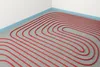 A close-up view of red underfloor heating pipes laid out in a pattern on a gray tiled floor, with a blue edge strip and white wall in the background. Náhľad obrázku produktu