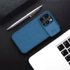 A blue iPhone case with a sliding camera cover is placed on a dark wooden surface next to a silver pen and a laptop. Náhľad obrázku produktu
