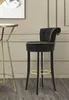 A black bar stool with gold trim is shown in a room with paneled walls and a wooden floor. A small gold table is partially visible to the left. Podgląd zdjęcia produktu