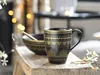 A black mug and bowl set with gold mandala patterns are displayed on a rustic wooden stool, with blurred bokeh lights in the background. Náhľad obrázku produktu