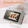 A vintage-style digital camera necklace is displayed diagonally against a soft, pinkish-white background with some decorative dried flowers. Produktbild-Vorschau