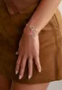 A close-up shot of a woman's arm wearing a silver charm bracelet with a pink charm, resting on a brown textured surface. Anteprima dell’immagine prodotto
