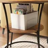 A close-up view of a round side table with a shelf, holding a white storage bin filled with books. Náhľad obrázku produktu