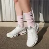 A person wearing white Nike Air Max Plus sneakers with pink socks, standing in front of a corrugated metal wall. Náhled obrázku produktu