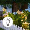 Solar watering can lights illuminate a garden at dusk, with a white picket fence in the foreground. Anteprima dell’immagine prodotto