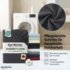 A folded fitted sheet is shown in the foreground, with a stack of other colored sheets behind it. A washing machine is visible in the background. Produktbild-Vorschau