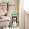 A narrow green side table with two shelves is placed next to a beige armchair. The table has a woven basket on the top shelf and two storage boxes on the bottom shelf. A glass of water and a book are on top of the table. The scene is set in a living room w Náhľad obrázku produktu