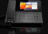 A dark-colored VoIP phone with a touchscreen display and handset, set against a black reflective surface and background. Produktbild-Vorschau