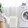 A white laundry bin with a textured pattern sits next to a washing machine, with a laundry basket filled with white towels in the foreground. Produktbild-Vorschau