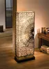 A tall, rectangular floor lamp with a woven rattan shade is illuminated in a room with wooden floors and a window in the background. Náhled obrázku produktu