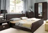 A bedroom scene featuring a dark wood bed, nightstand, dresser, and wardrobe with a mirror. Podgląd zdjęcia produktu
