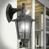 A black outdoor wall lantern is mounted on a white wall, with a modern building in the background. Podgląd zdjęcia produktu
