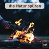 A person is adding a log to a campfire in a dark, outdoor setting. Produktbild-Vorschau