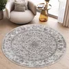 A round, vintage-style rug with a distressed pattern in shades of gray and white is placed on a wooden floor in a living room setting. A modern, light-colored armchair with cushions sits to the left of the rug, and a small, gold-toned side table with a boo