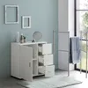 A white bathroom cabinet with drawers and a door, placed on a light blue wall next to a window and a towel rack. Náhled obrázku produktu